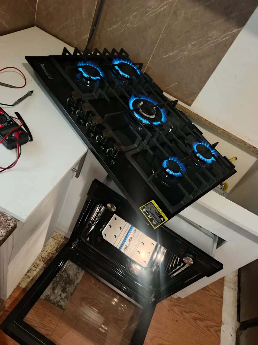 Encimera de gas negra con cinco hornillas encendidas de llama azul, con un horno abierto debajo y cables de medición a la izquierda