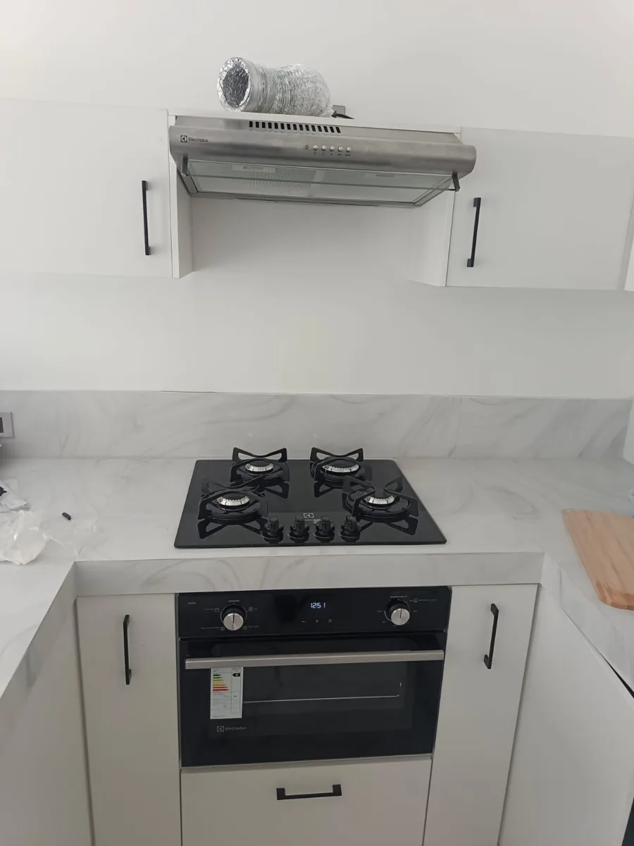 Encimera de gas negra de cuatro hornillas y horno negro debajo, con campana extractora arriba, en una cocina blanca