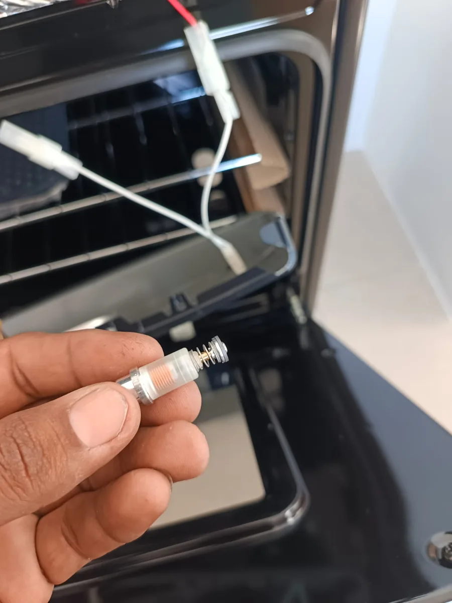 Mano sosteniendo una pieza pequeña con resorte frente a un horno con la puerta abierta y un cable blanco